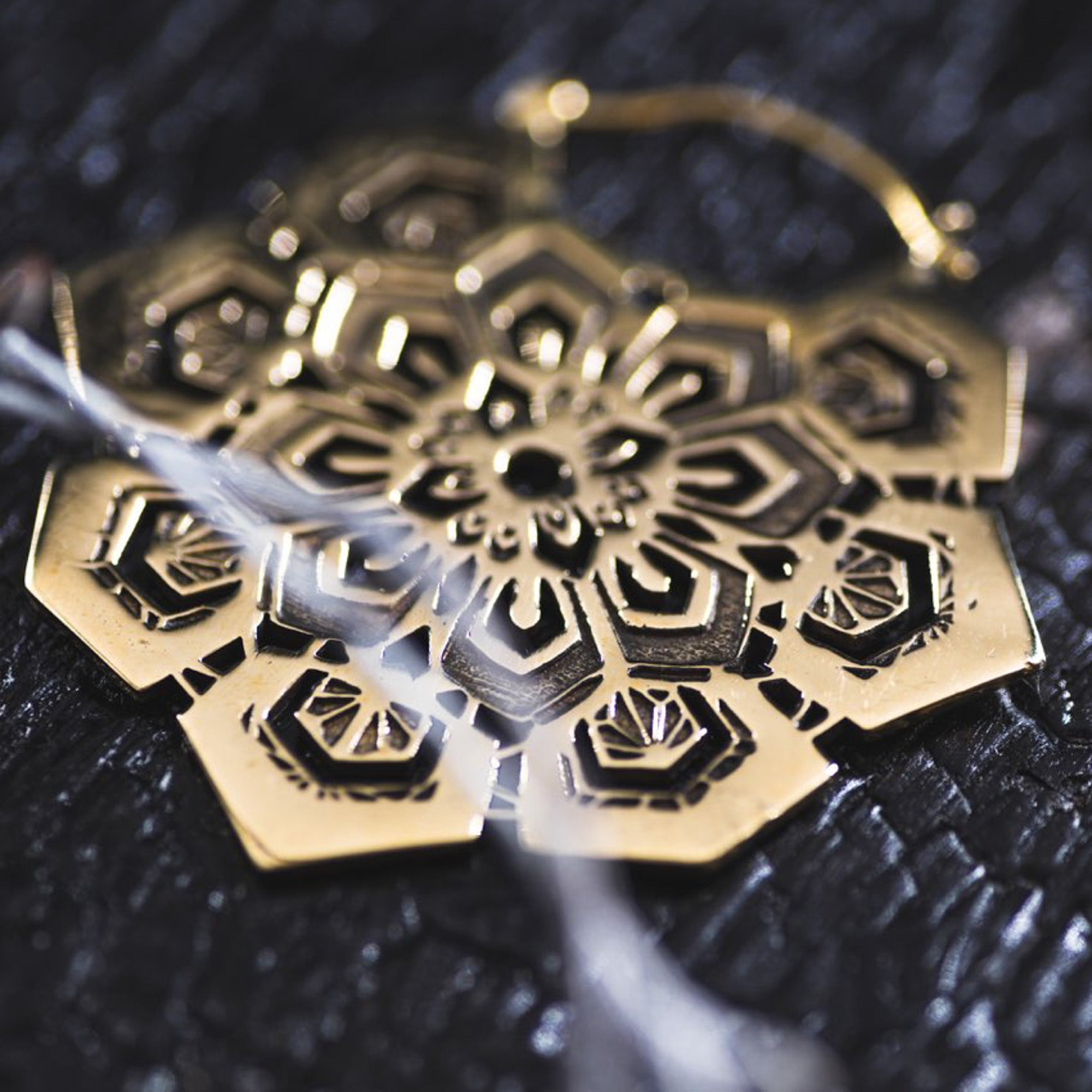 A close-up of a detailed ASA Oversized Asa No Ha Mandala Disc Earring in Gold by Kaya Movement lying on a textured dark surface. The design features intricate geometric patterns and cutouts, reminiscent of ethnic statement earrings, surrounding a central flower-like shape. A blurred white feather is partially visible in the foreground.
